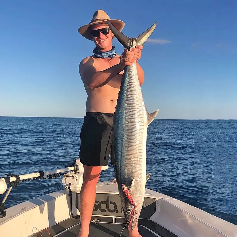 Fisher with a Spanish mackerel, standing on a boat deck with expansive ocean waves behind him.