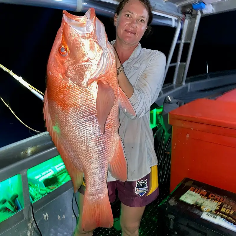 Angler aboard a boat cradling a large red emperor fish, with dim night lighting illuminating the catch.