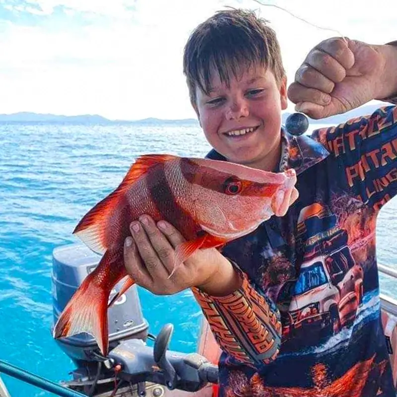 Smiling young angler holding a red emperor fish aboard a boat, with turquoise sea in the background.