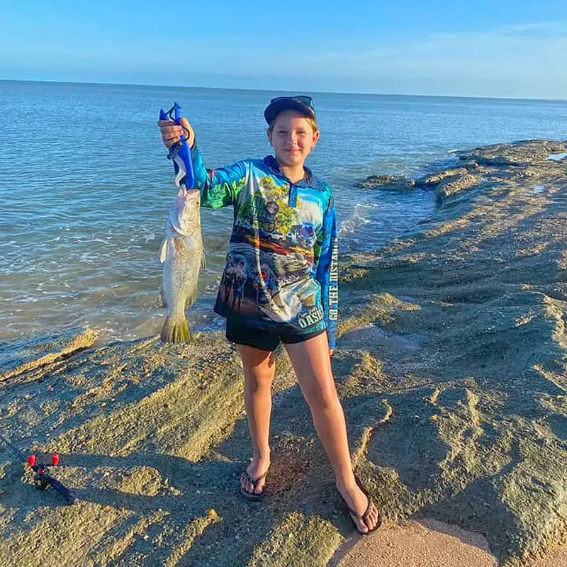 Fisher standing on shoreline, holding a golden trevally with clear blue ocean in the background.