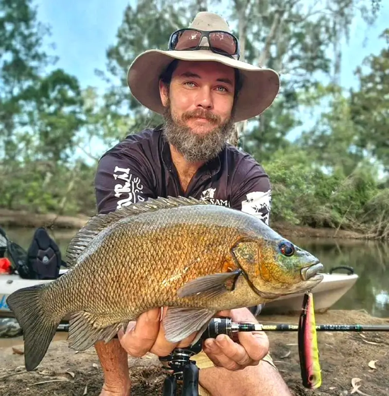 Fisher sitting on a riverbank, proudly displaying a golden perch, surrounded by bushland.