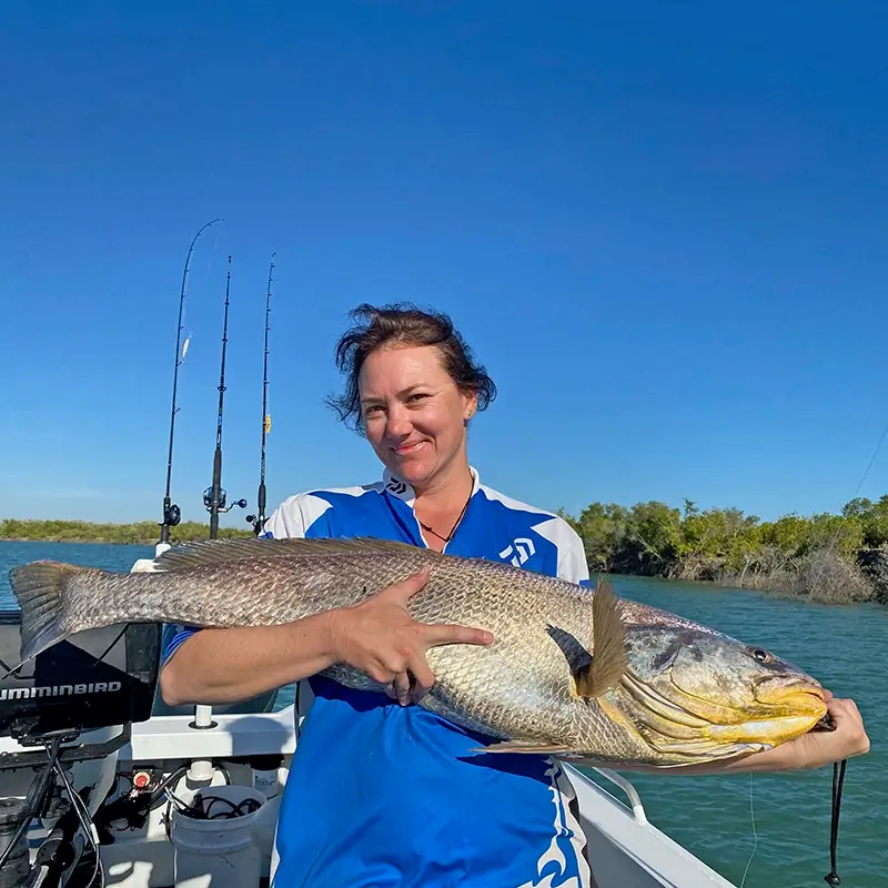 Woman on a boat holding a black jewfish, with calm waters and distant trees under a blue sky.