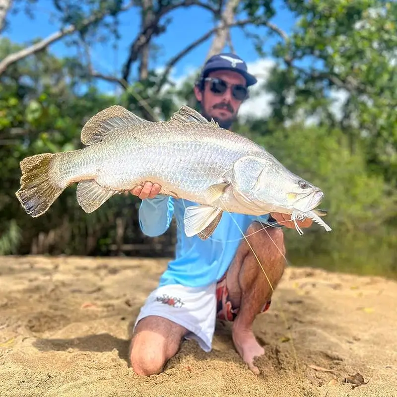 Angler kneeling on a sandy riverbank, holding a large, gleaming barramundi, with lush greenery and clear blue skies in the background.