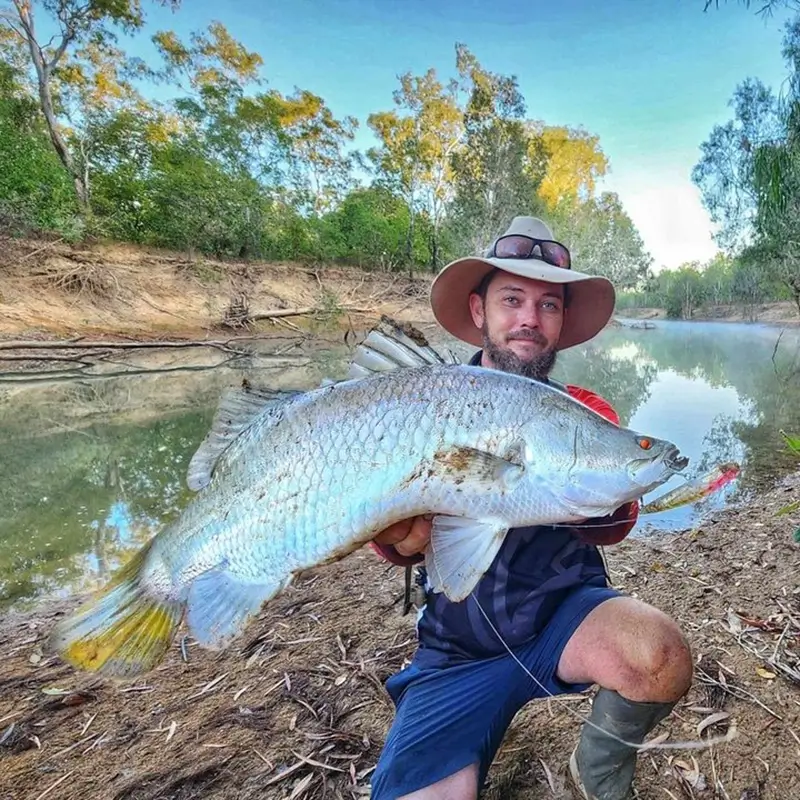 Young angler proudly displaying a freshly caught fish on a sunny day by the water’s edge, capturing the joy of a successful fishing trip at GBR Tackle & Bait Mareeba.