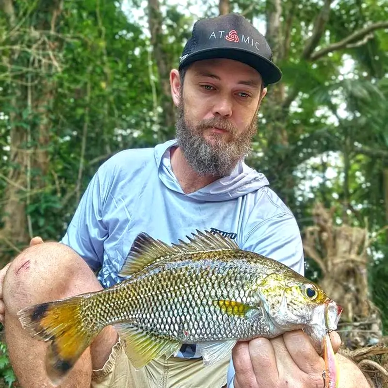 Man holding a yellowfin bream, caught along a lush riverside.