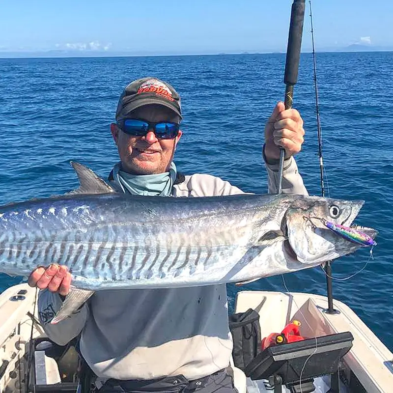 Fisher presenting a Spanish mackerel on a boat, against a deep blue ocean backdrop.