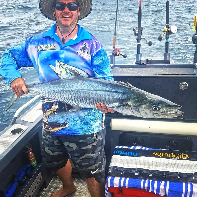 Angler standing on a boat, holding a freshly caught Spanish mackerel, with rods and ocean waves behind.