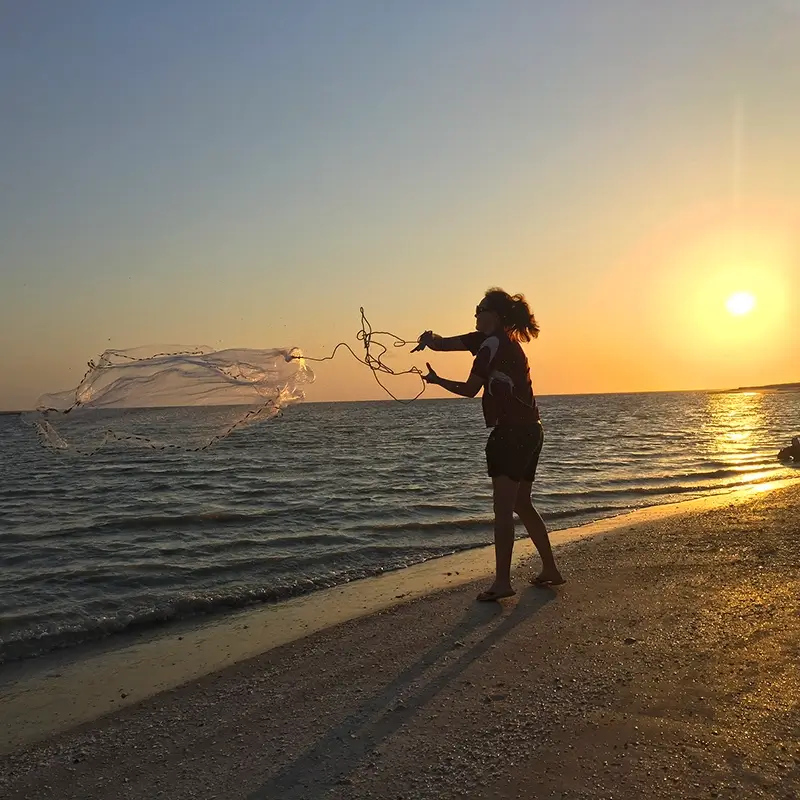 Silhouette of a person casting a fishing net along a sandy beach, with the setting sun glowing over the sea.