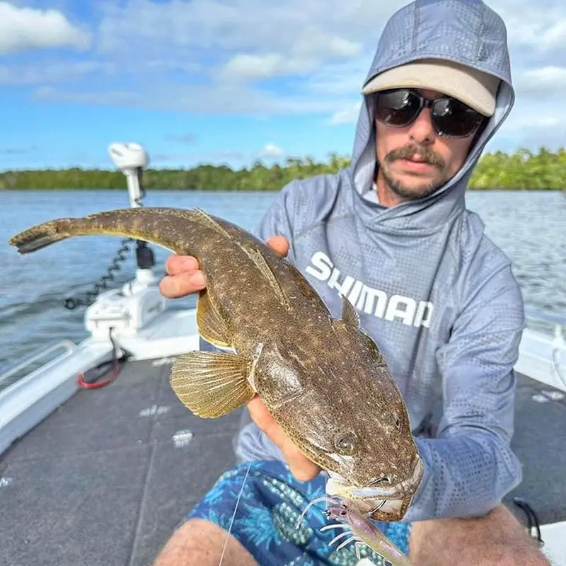 Angler displaying a dusky flathead, seated on a boat with low mangroves in the distance.