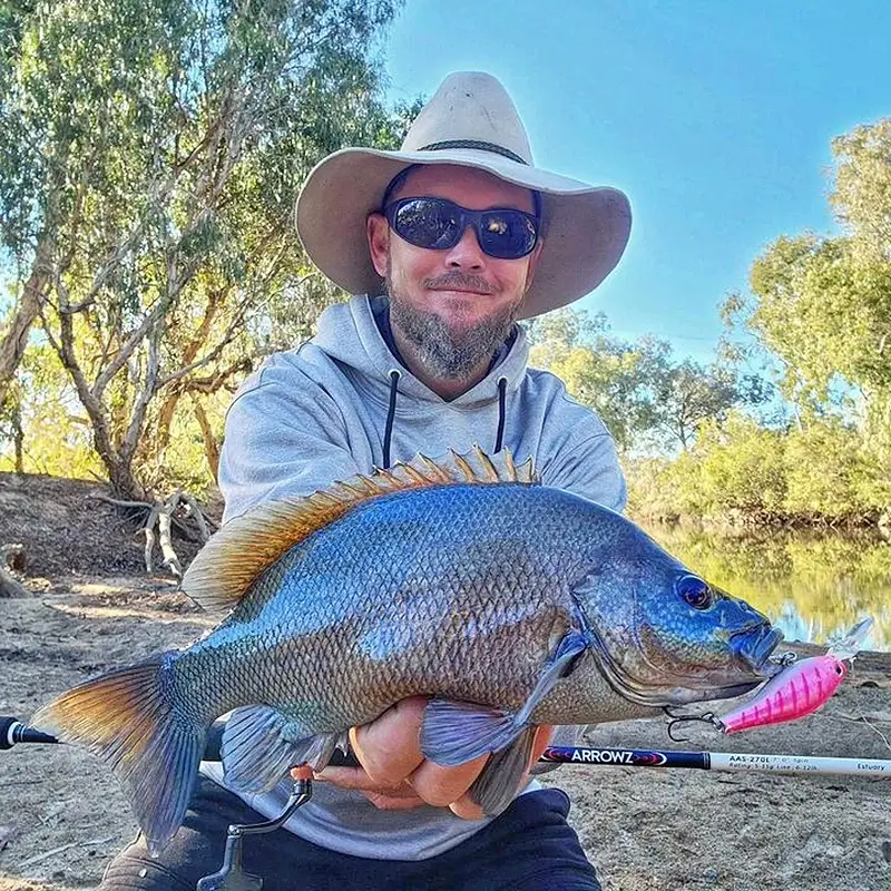 Fisher standing on a riverbank, holding a blue salmon with sunlit trees in the background.