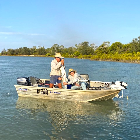 Two people fishing in a tinny boat on a calm river, with greenery along the banks.