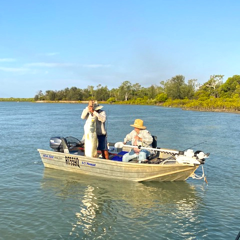 Two fishers seated in a tinny boat, capturing a moment of landing a fish in a wide, peaceful river.