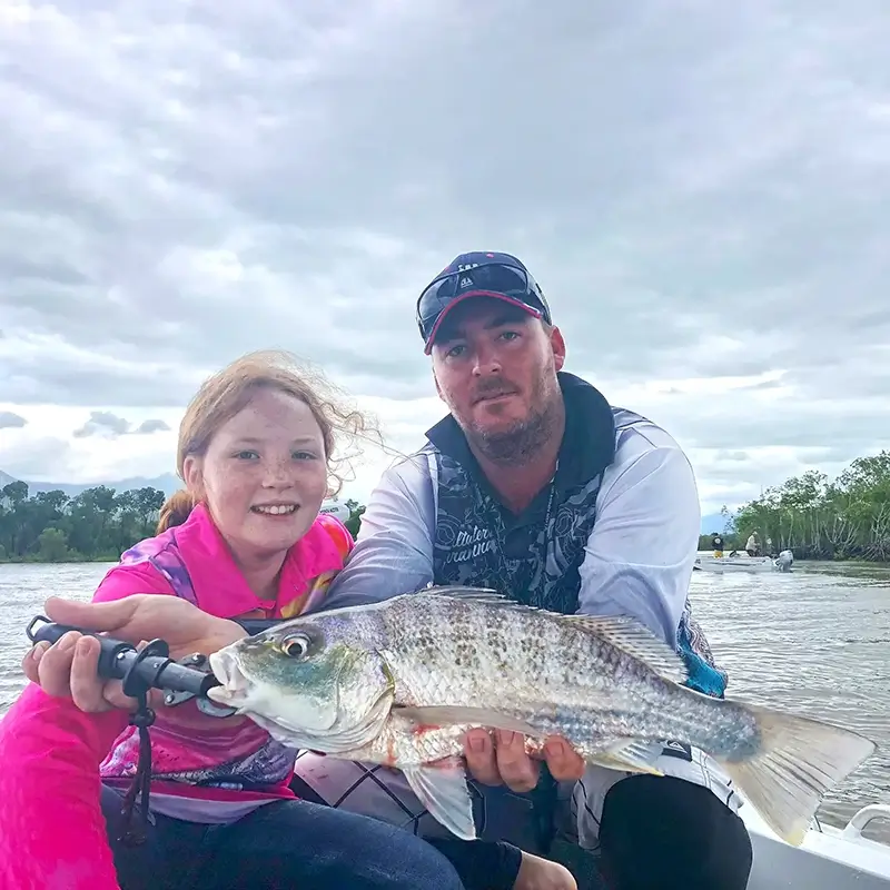 Father and daughter holding a freshly caught grunter together on a boat, with cloudy skies and mangroves in the background.