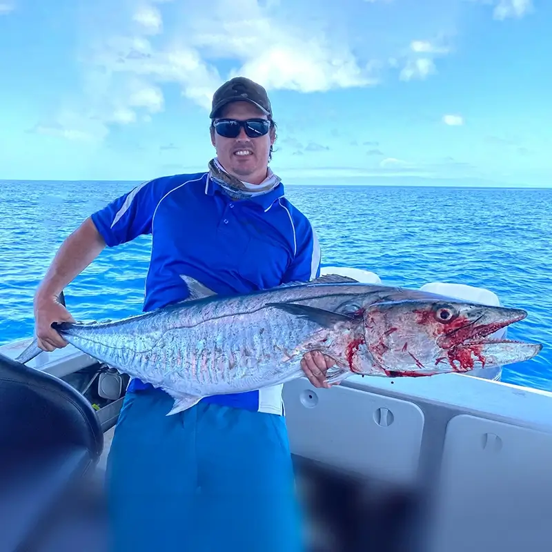 Fisher standing on a boat, proudly displaying a large Spanish mackerel against the clear blue ocean backdrop.