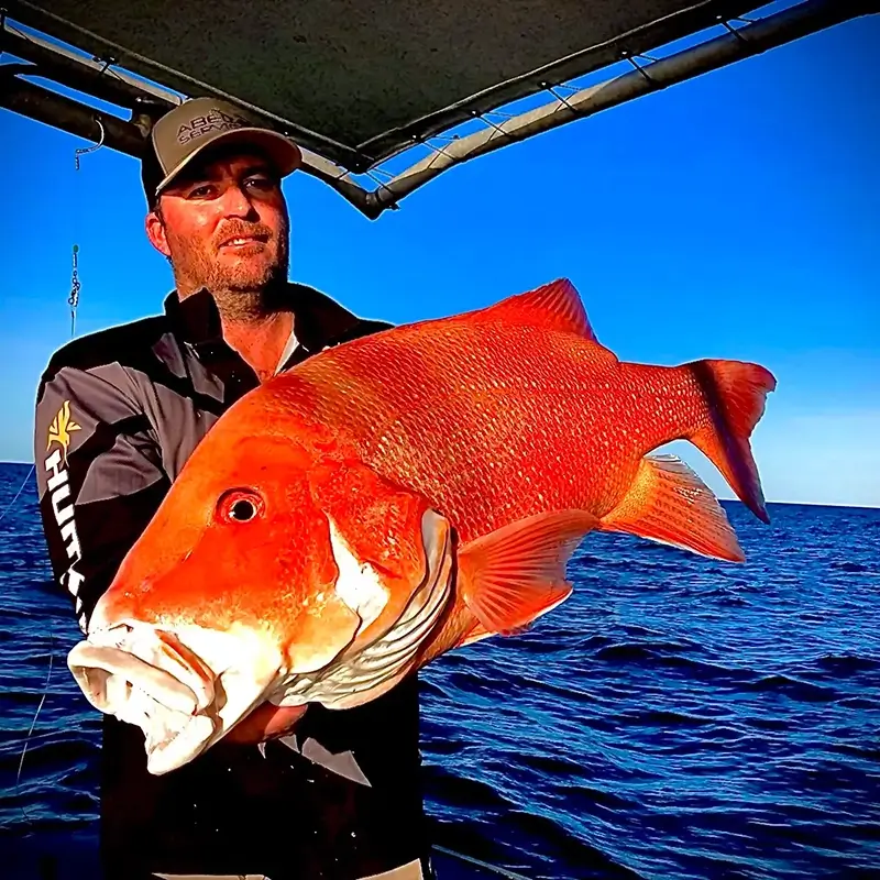 Mareeba fisher, Trevor Roy on a boat showcasing a vivid red emperor fish, with deep blue ocean waters behind him.