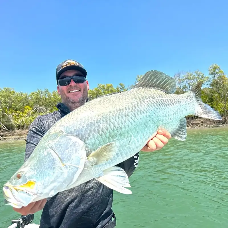 Angler holding a massive silver barramundi, standing in a boat with a river and dense foliage behind him.